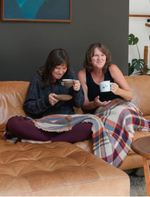 Two women sitting on a brown couch in a living room drinking tea.
