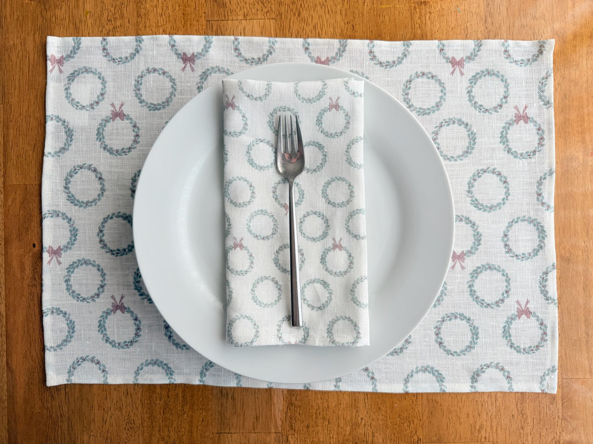A place setting on a wooden table featuring a white linen placemat with a repeating wreath and bow pattern, a white ceramic plate, a folded white napkin with a matching wreath print, and a silver fork.
