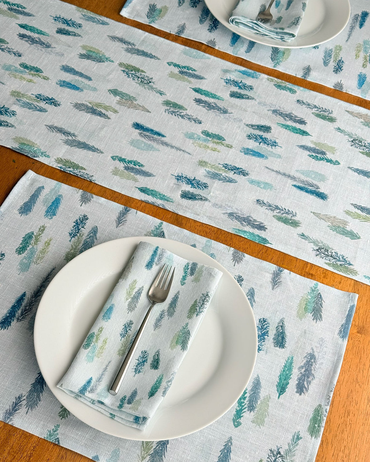 A set of light blue table linens featuring a repeating pattern of blue and green magical pine trees, styled on a wooden table with white plates. matching placemats and folded napkins.