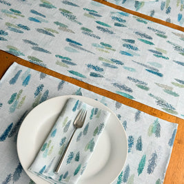 A set of light blue table linens featuring a repeating pattern of blue and green magical pine trees, styled on a wooden table with white plates. matching placemats and folded napkins.