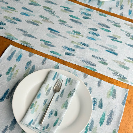 A set of light blue table linens featuring a repeating pattern of blue and green magical pine trees, styled on a wooden table with white plates. matching placemats and folded napkins.