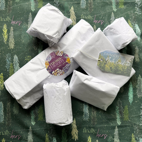Several white paper-wrapped packages arranged on a dark green forest-patterned fabric background. One package has a moss + bloom sticker. A hanging tag reads rooted in North Carolina.