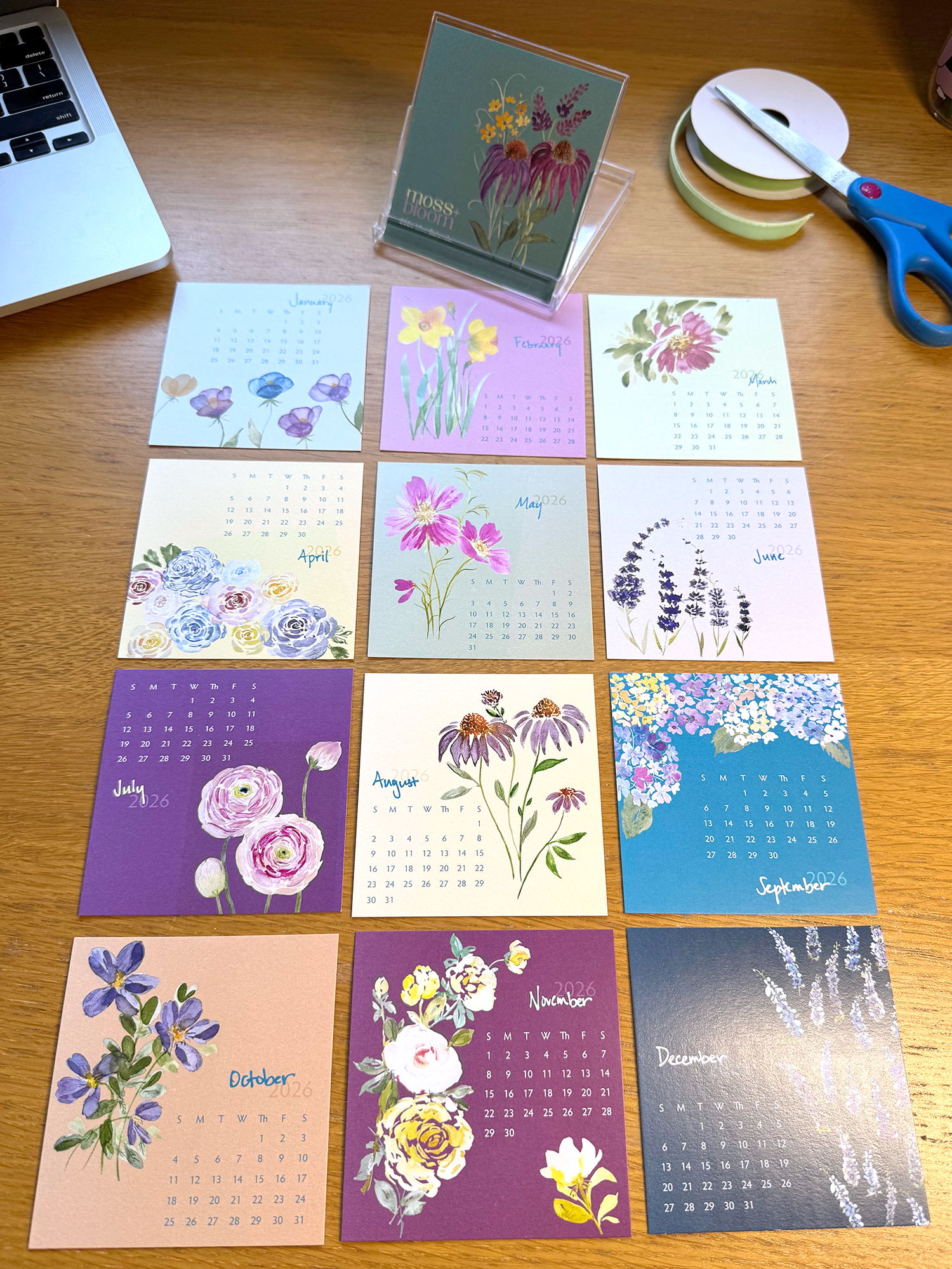 Set of floral-themed calendars on a wooden surface with a laptop, scissors, and tape dispenser.