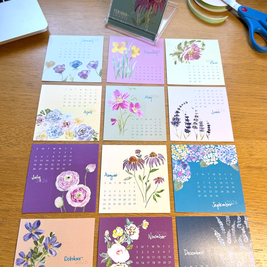 Set of floral-themed calendars on a wooden surface with a laptop, scissors, and tape dispenser.