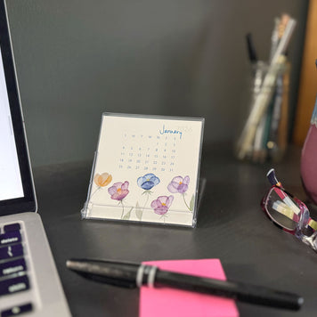 Desk setup with a laptop, calendar, pen, and glasses on a dark surface.